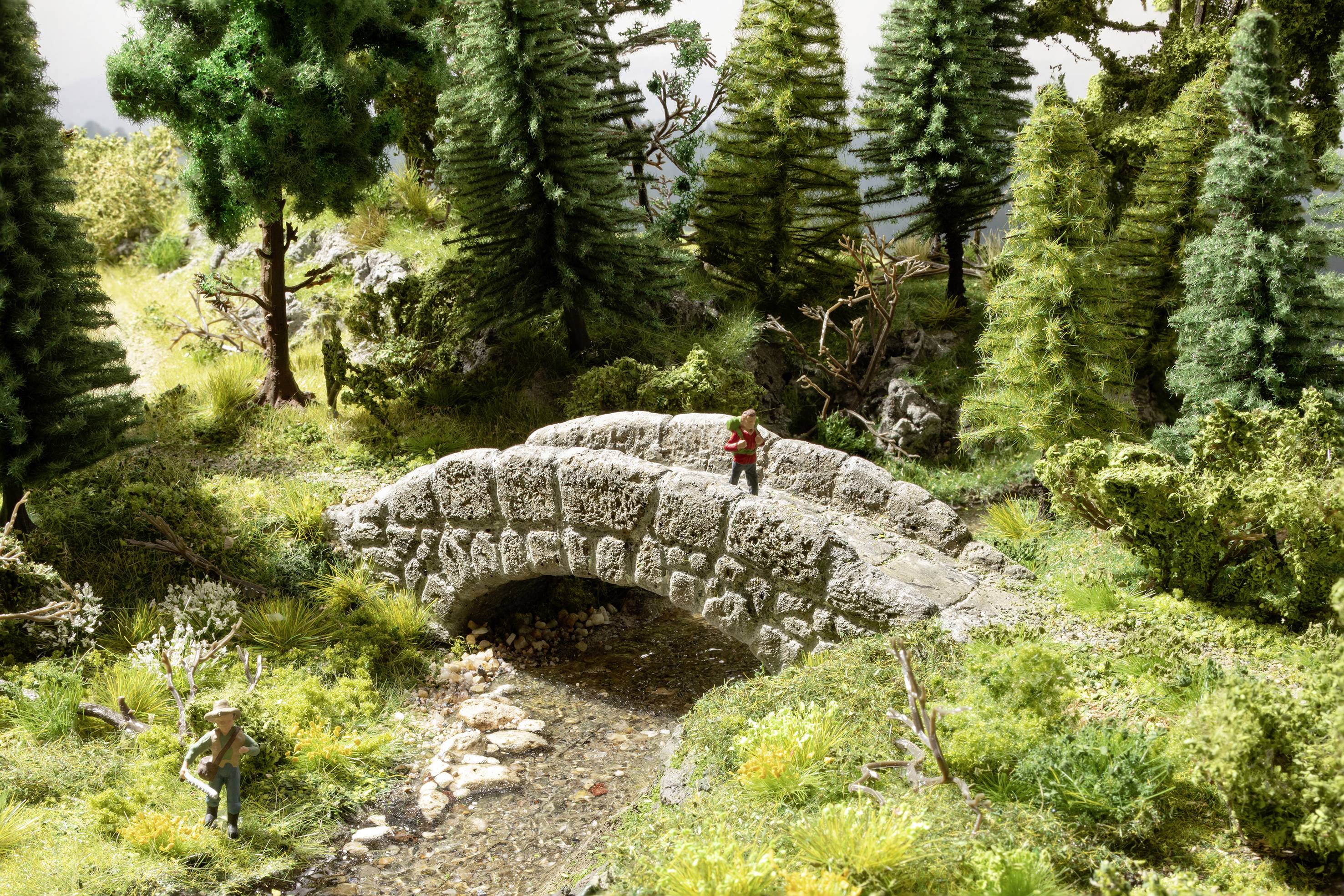 Ein kleiner Steinbogen überquert einen Bach in einem waldreichen Gebiet. Zwei Modellfiguren wandern darauf zu.