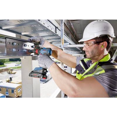 Ein Bauarbeiter mit Schutzhelm und Brille befestigt mit einer Bohrmaschine eine Metallplatte in einem Gebäude.