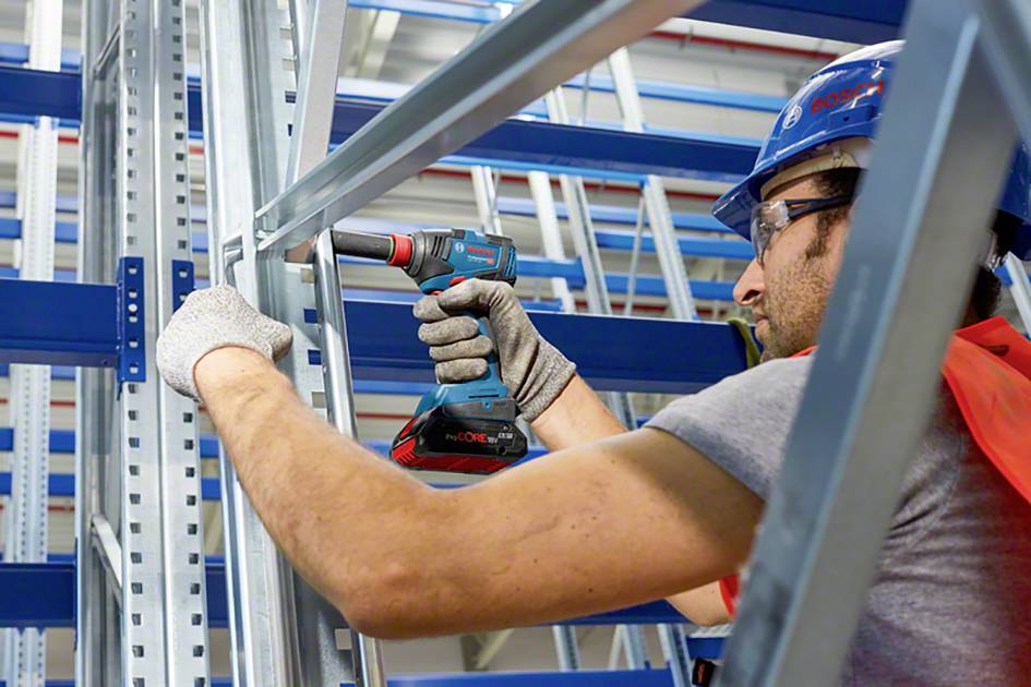 Eine Person in Schutzhelm und Handschuhen montiert Metallregale mit einem Akku-Bohrschrauber in einem Lagerraum.