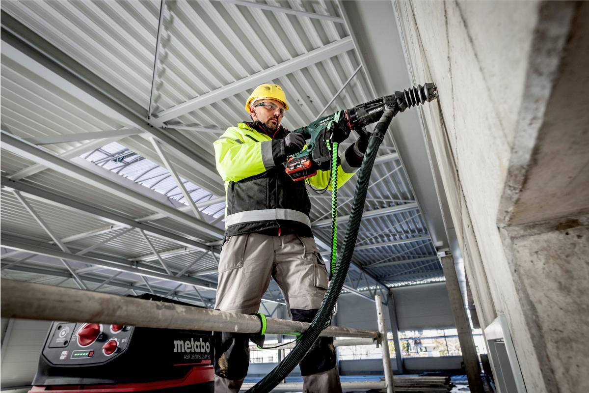 Ein Bauarbeiter mit Schutzhelm und Arbeitskleidung benutzt einen Bohrhammer an einer Betonwand in einer großen Halle.