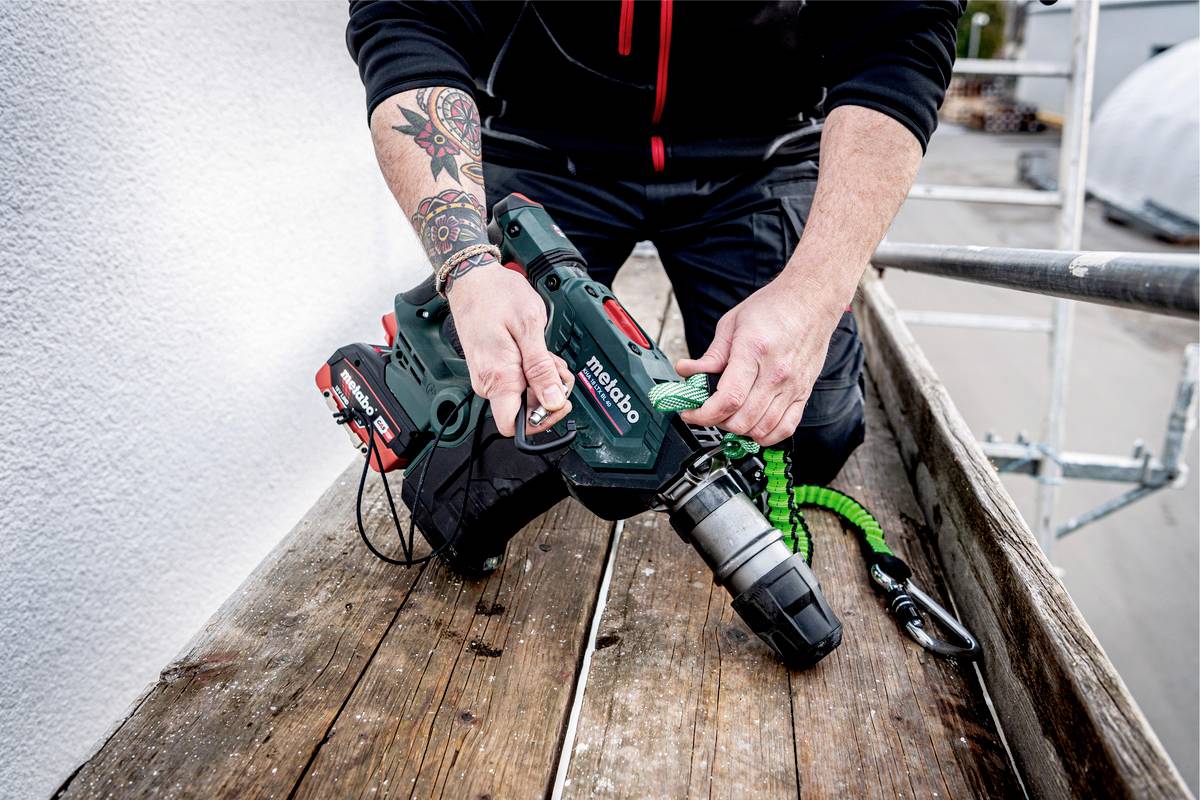 Eine Person hält eine grüne Bohrmaschine auf einem Holzboden. Die Bohrmaschine wird gesichert, und die Person hat Tattoos auf dem Arm.