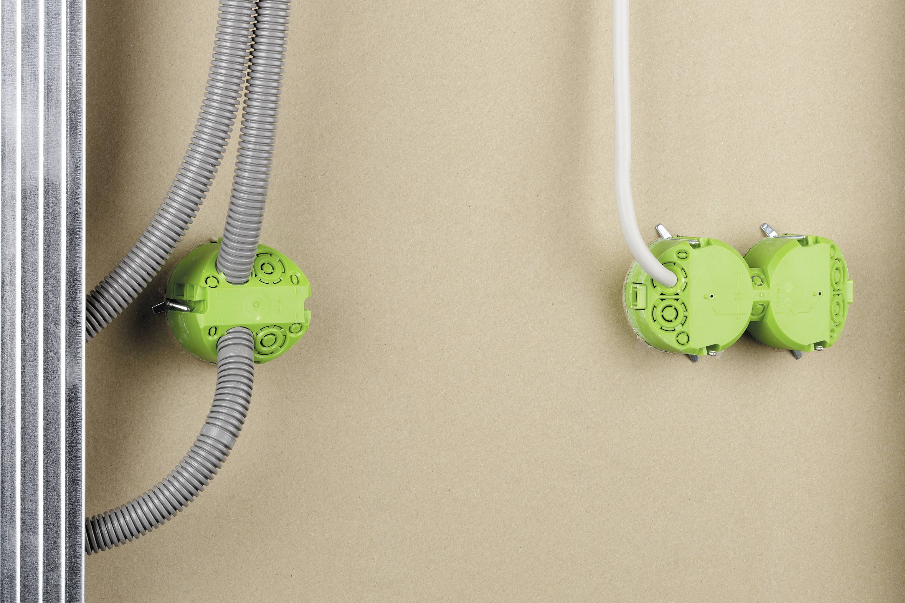 Zwei grüne Elektroanschlussdosen mit grauen und weißen Kabeln an einer beigen Wand, teilweise von einem Metallrahmen verdeckt.