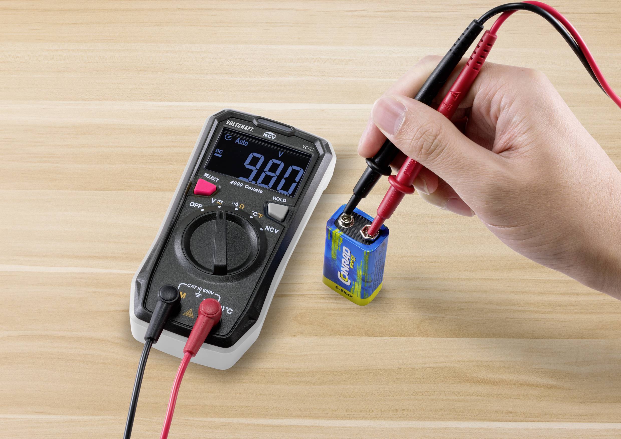 Eine Person misst die Spannung einer 9-Volt-Batterie mit einem Digitalmultimeter. Das Display zeigt 9,8 Volt an.