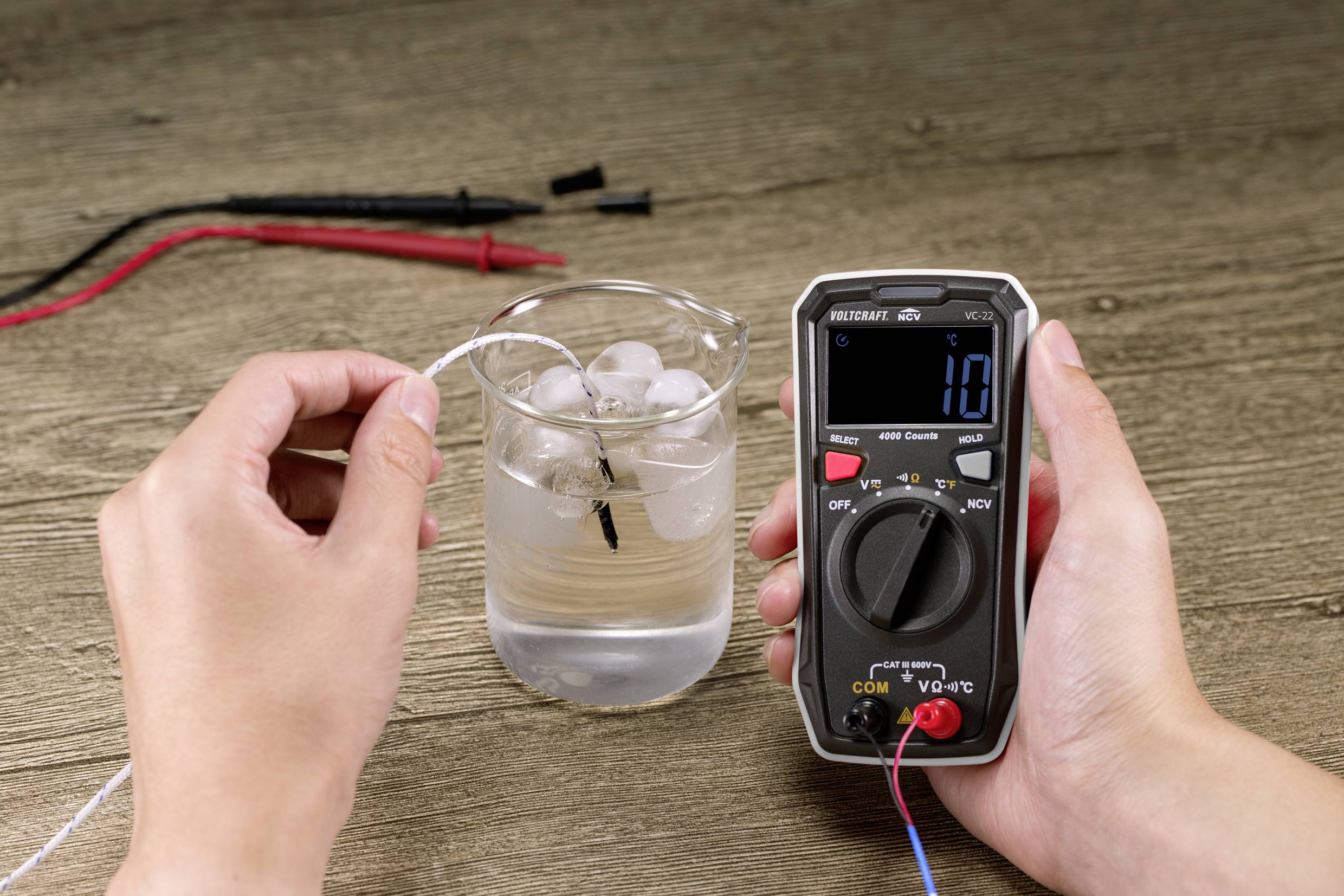 Eine Person misst mit einem Multimeter die elektrische Leitfähigkeit von Wasser in einem Glas mit Eiswürfeln. Die Anzeige zeigt '10' an.