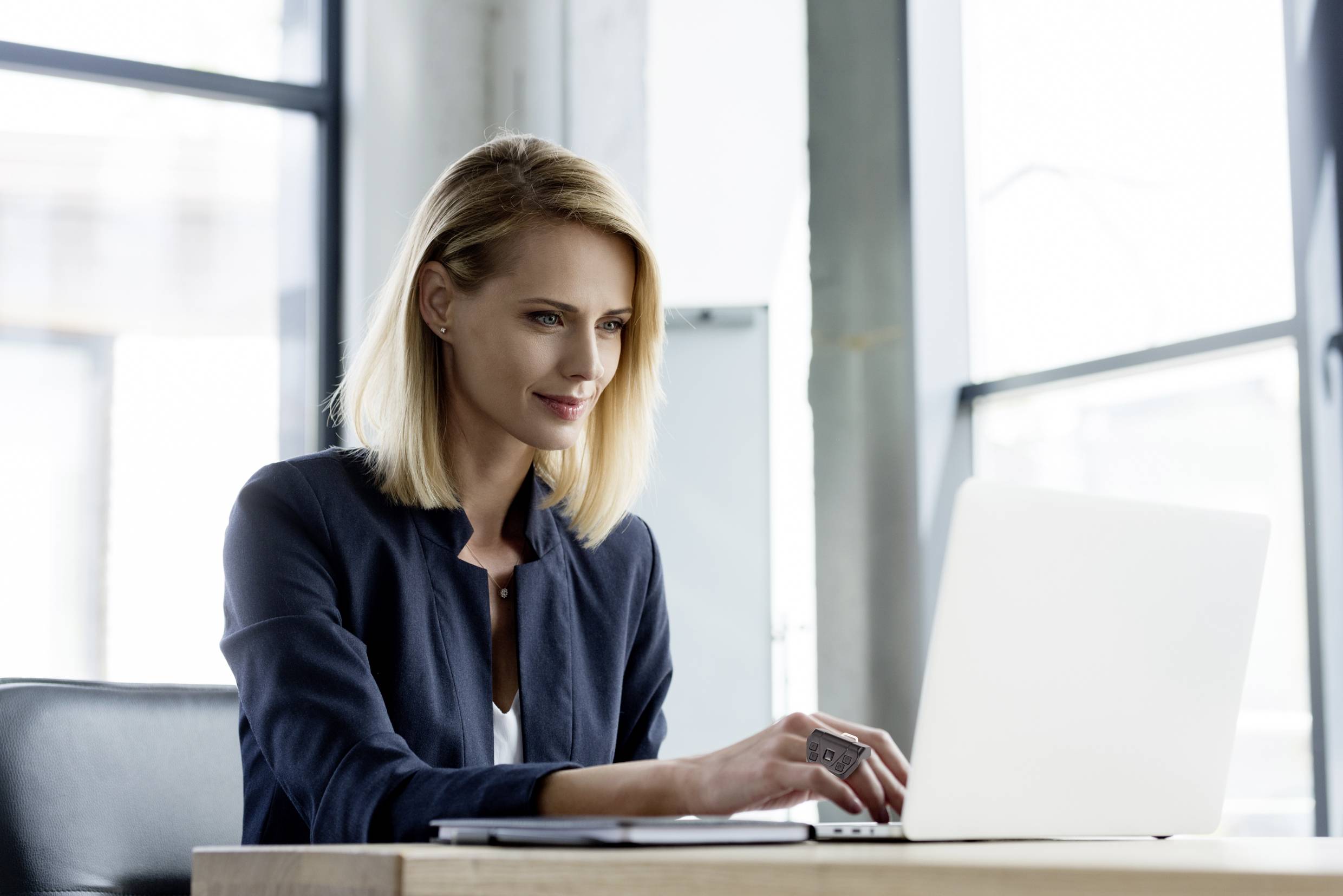 Eine Frau mit blondem Haar sitzt an einem Tisch und arbeitet konzentriert an einem Laptop. Hintergrund zeigt große Fenster.