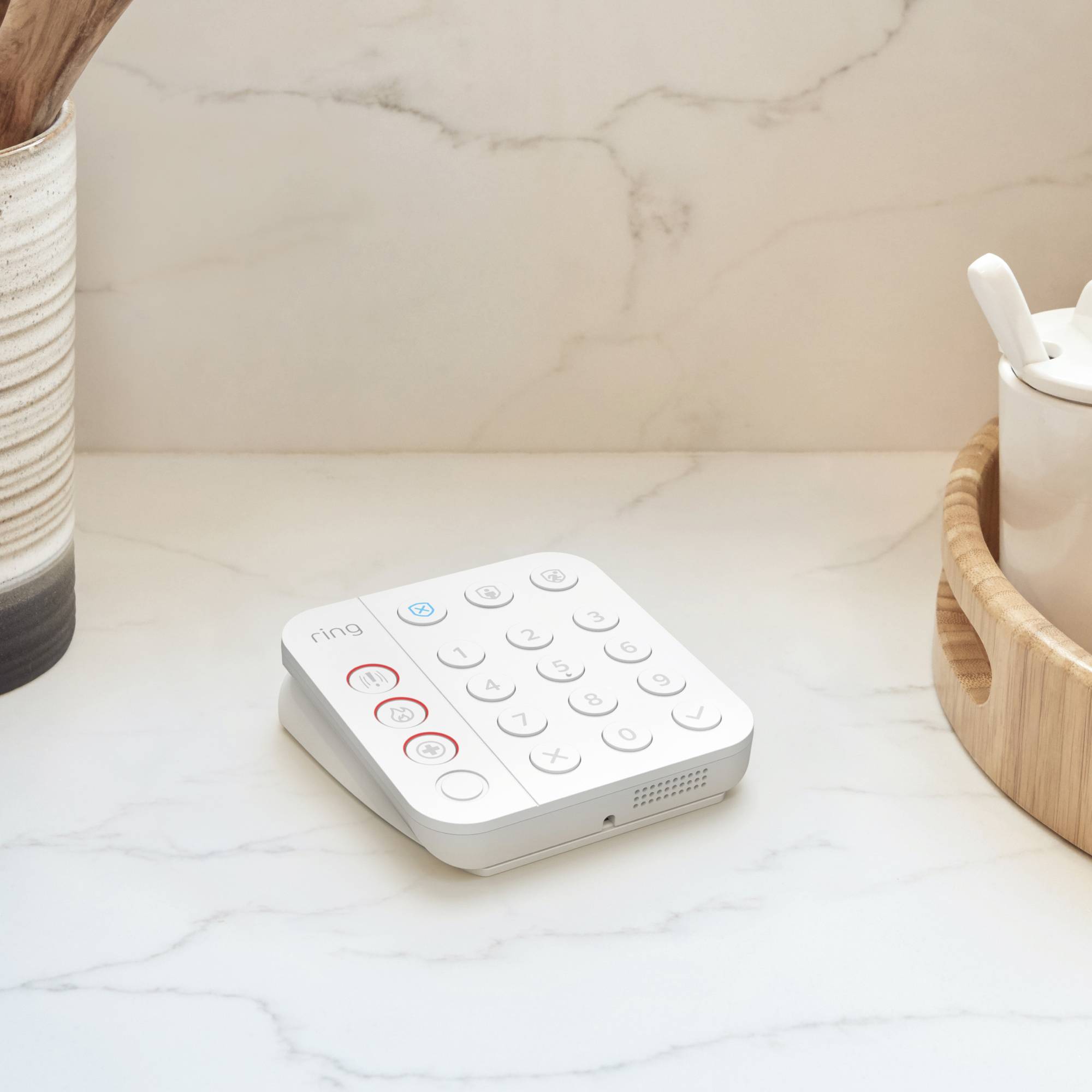 Ein weißes Smart-Home-Alarmtastenfeld mit Tasten und Symbolen steht auf einer Marmoroberfläche, neben einem Holztablett.