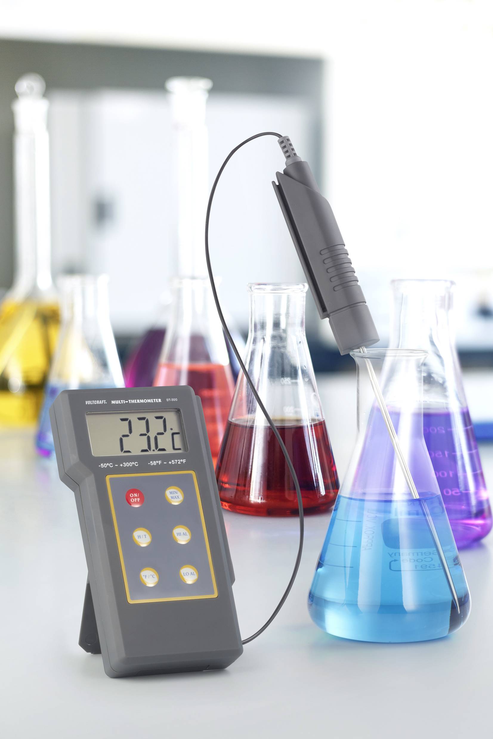 Laborthermometer misst 23,2°C in einer blauen Flüssigkeit in einem Erlenmeyerkolben. Andere Kolben mit farbigen Flüssigkeiten im Hintergrund.