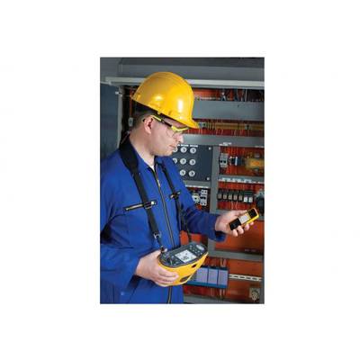Ein Elektriker in blauer Arbeitskleidung und gelbem Schutzhelm überprüft Messgeräte vor einem Schaltschrank mit elektrischen Komponenten.