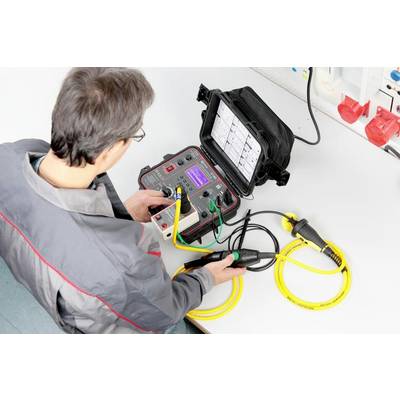 Eine Person bedient ein elektrisches Testgerät an einem Tisch. Das Gerät hat gelbe Kabel und ein Display, das Messdaten anzeigt.