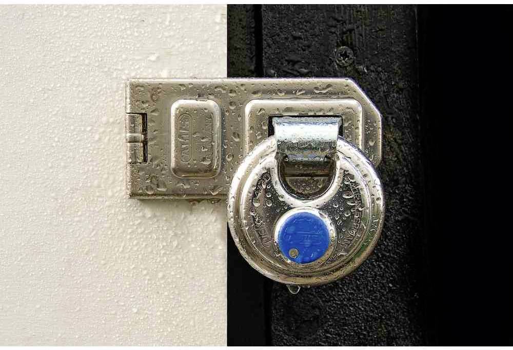 Ein silbernes Vorhängeschloss sichert eine schwarze Tür, befestigt an einem metallischen Scharnier auf einer hellen Oberfläche. Wassertropfen sind sichtbar.