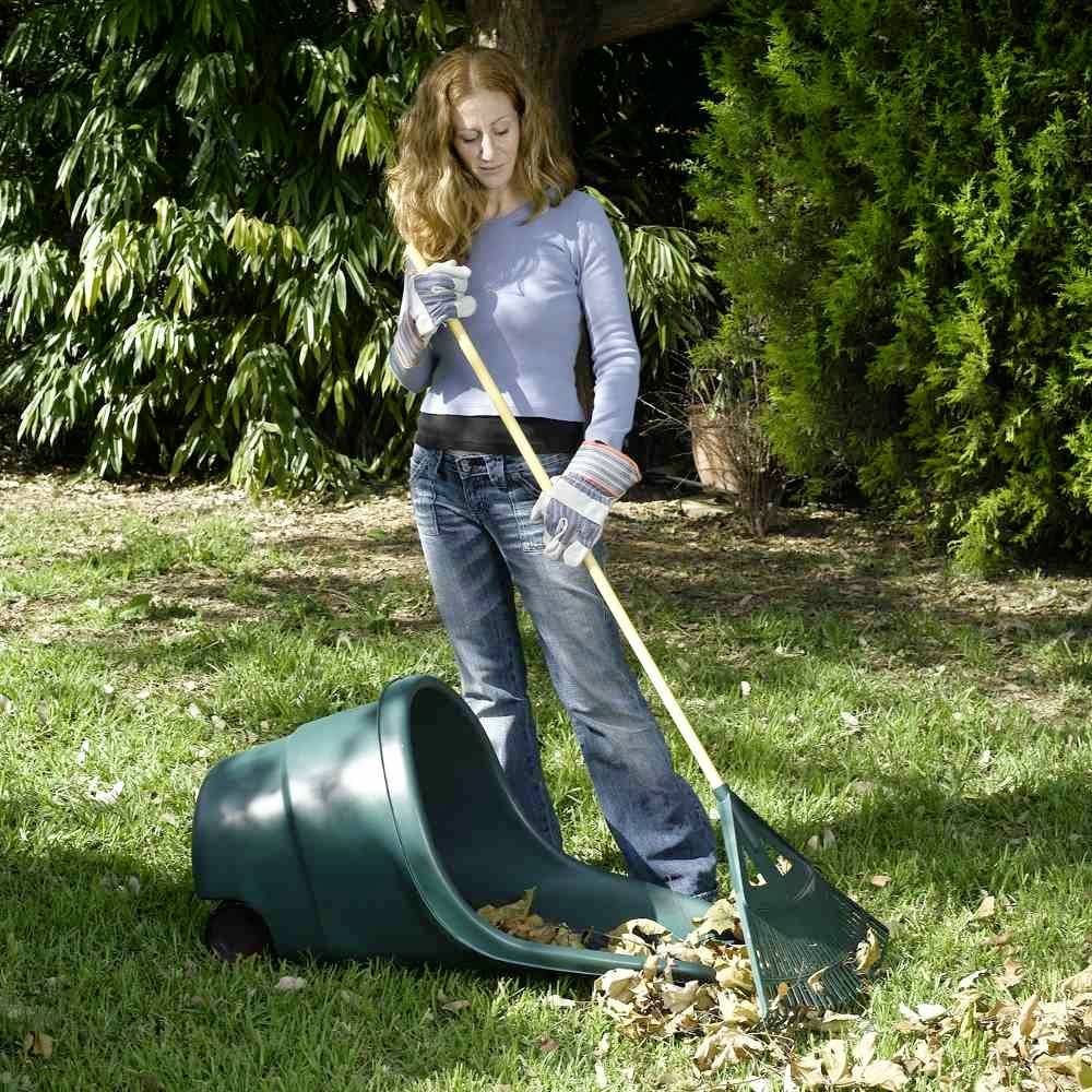 Eine Person harkt Herbstblätter in einem Garten zusammen und sammelt sie in einem grünen Behälter. Umgeben von üppigem Grün.