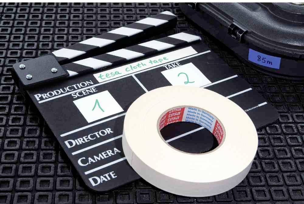 Klappe mit den Beschriftungen 'Scene 1' und 'Take 2' neben einer Rolle Klebeband auf einem strukturierten Untergrund.