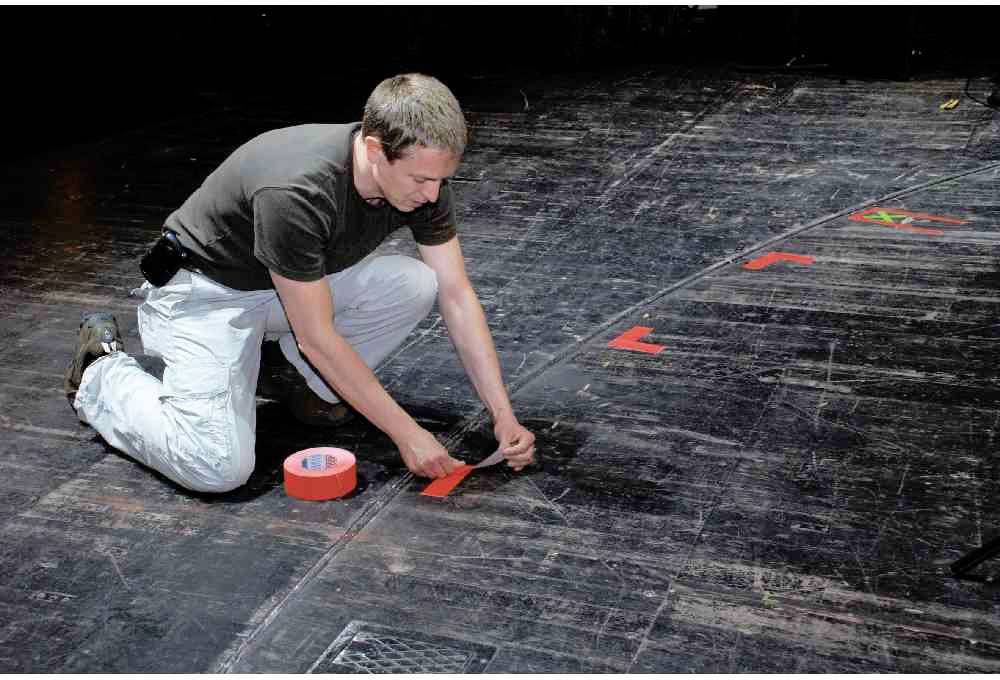 Ein Mann kniet auf einer Bühne und klebt rotes Klebeband in Form von Markierungen auf den Boden. Er konzentriert sich auf die genaue Platzierung.