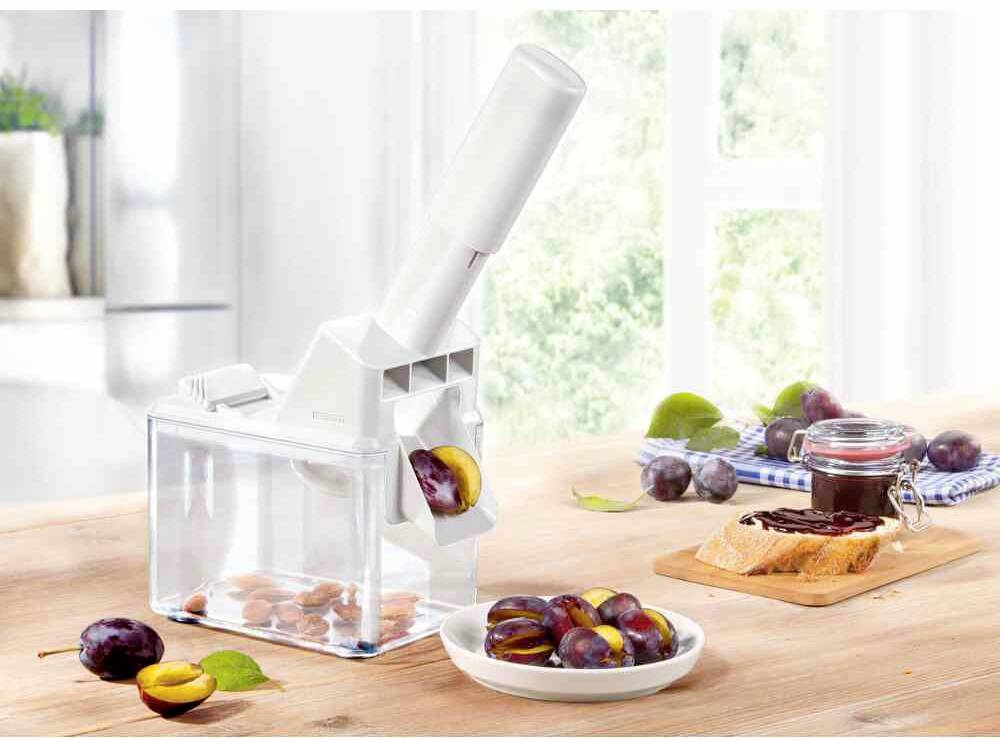 Ein Pflaumenentkerner auf einem Tisch mit Schale voller entkernter Pflaumen und einem Toast mit Marmelade im Hintergrund.