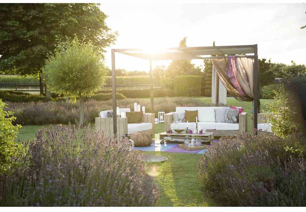 Eine gemütliche Outdoor-Lounge im Garten bei Sonnenuntergang, mit Sofas unter einer Pergola, umgeben von Lavendelbeeten und Kerzen.