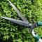 Eine Schere schneidet eine grüne Hecke. Die scharfen Klingen durchtrennen präzise die Blätter, während sie die Hecke formen.