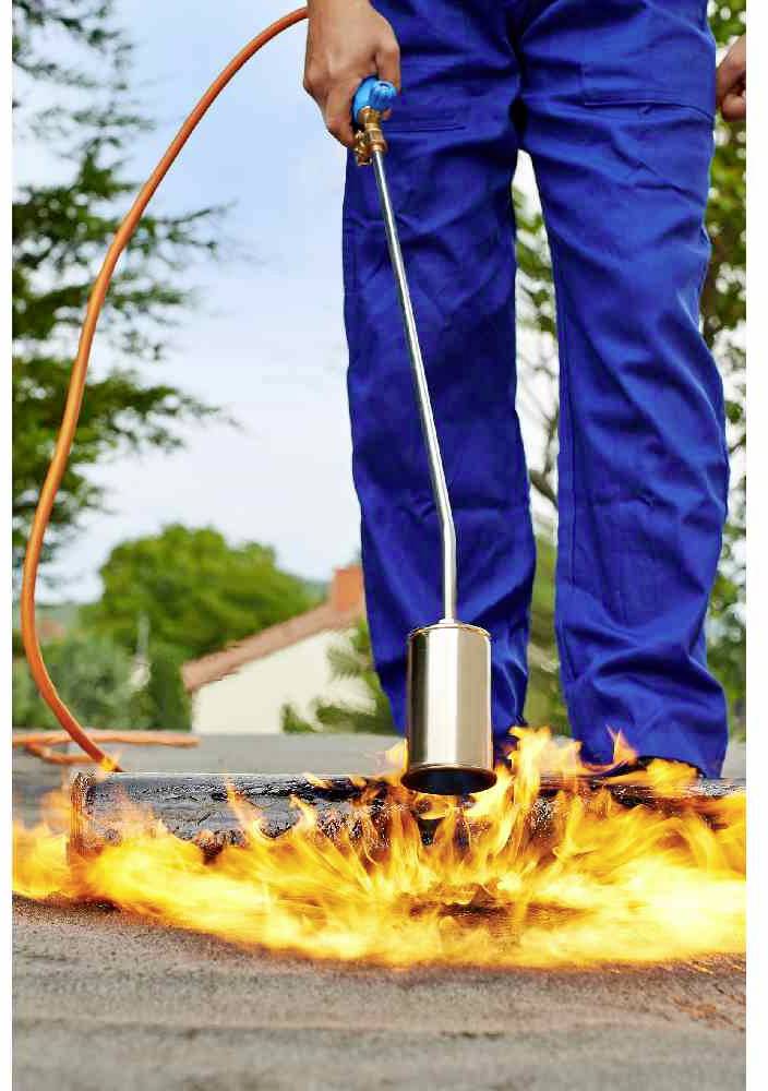 Ein Arbeiter in blauer Arbeitskleidung glüht mit einem Gasbrenner Bitumen auf einem flachen Dach. Flammen sind sichtbar.