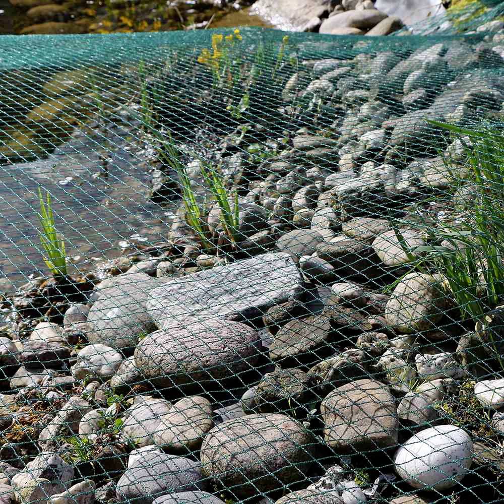 Steiniger Uferbereich eines Flusses, abgedeckt mit grünem Netz zum Schutz vor Erosion; sprießende Gräser zwischen den Steinen.