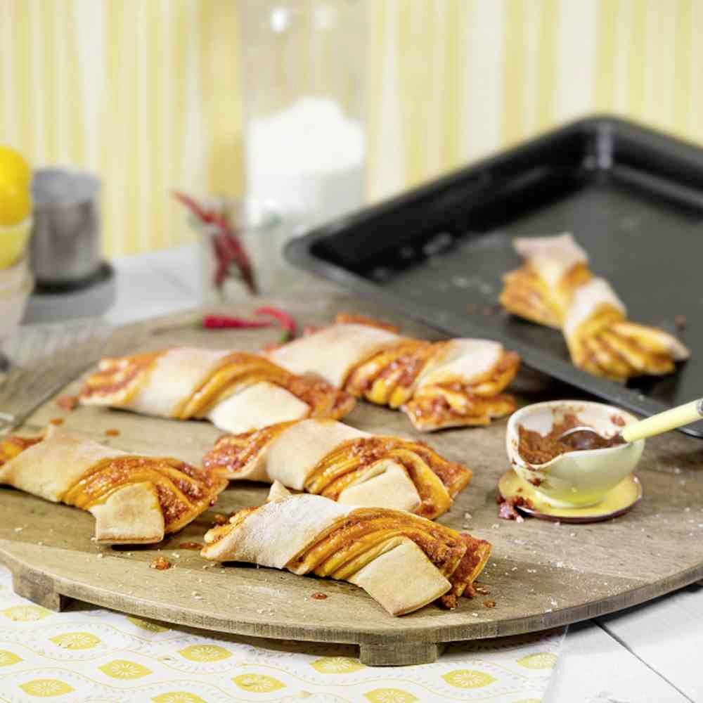 Eine hölzerne Platte zeigt mehrere gefüllte Teigtaschen mit Tomatensauce und Käse, im Hintergrund ein Tablett mit einer weiteren Portion.