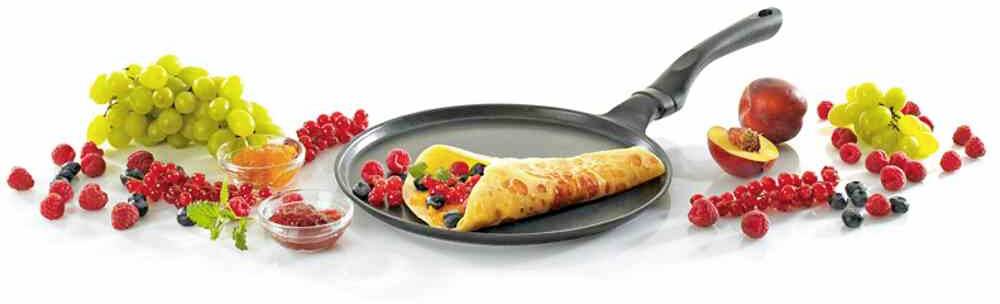 Ein gefüllter Crêpe liegt in einer Pfanne, umgeben von Trauben, Himbeeren, Pfirsichen und Honig, die eine bunte Frühstücksszene zeigen.