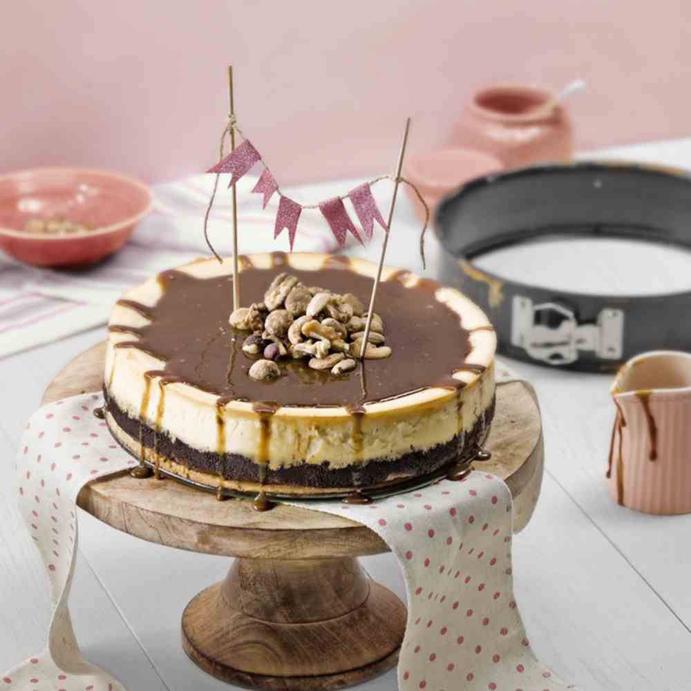 Kuchen auf Holzplatte, dekoriert mit Nüssen und kleiner Wimpelkette. Im Hintergrund rosa Geschirr und Springform.