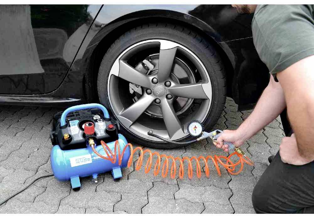 'Person pumpt Autoreifen auf, verbunden mit einem blauen Kompressor und orangefarbenem Schlauch, auf einer gepflasterten Fläche.'