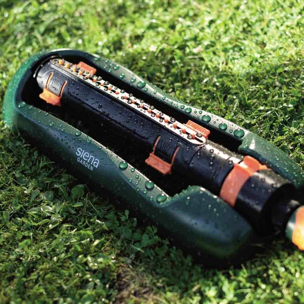 Ein grüner Rasensprinkler auf Gras, bewässert den Rasen. Der Sprinkler ist schwarz mit orangefarbenen Akzenten und Wassertropfen darauf.