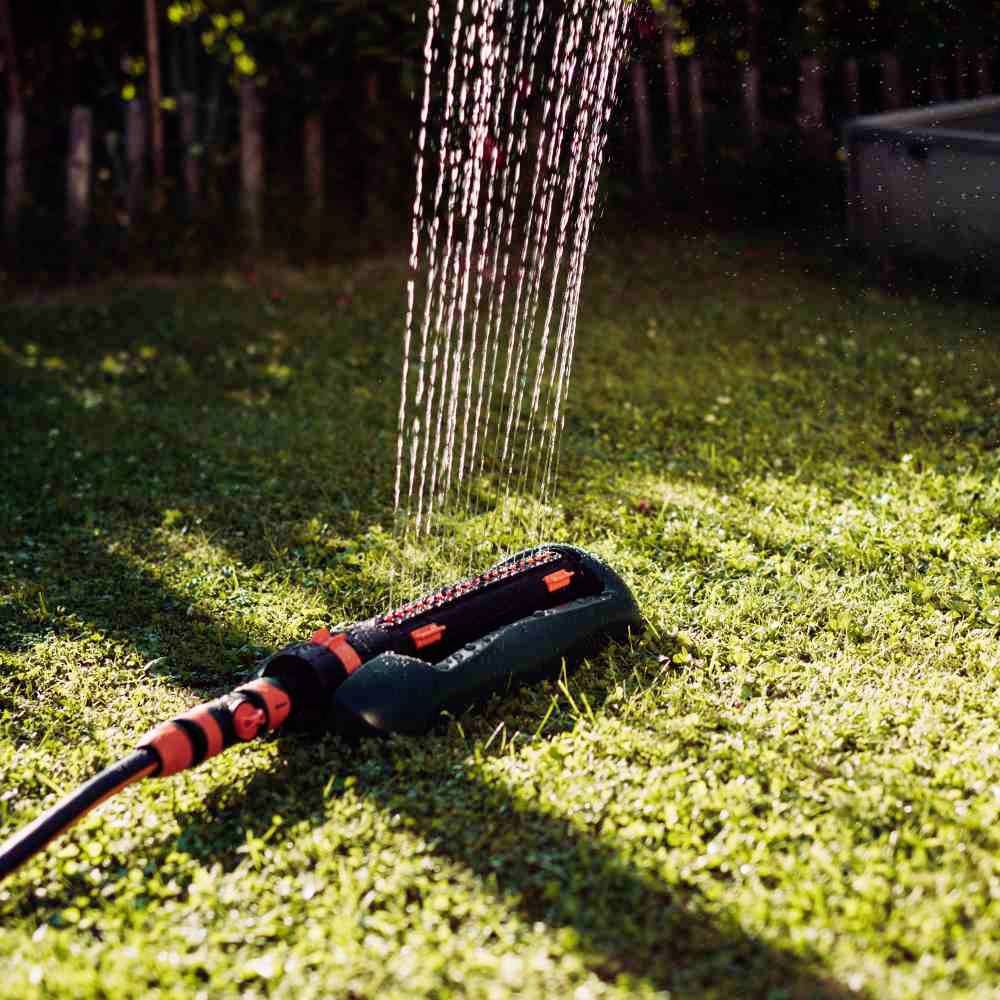 Ein Gartensprinkler bewässert einen sonnigen Rasen, wobei Sonnenstrahlen durch das Wasser schimmern.