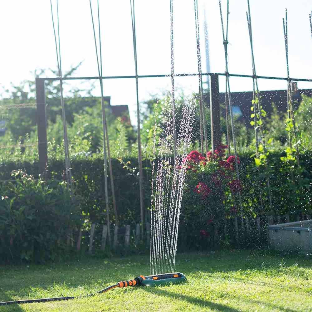 Ein Gartensprinkler bewässert einen grünen Rasen neben blühenden Sträuchern und Pflanzenstützen bei Sonnenschein in einem Garten.