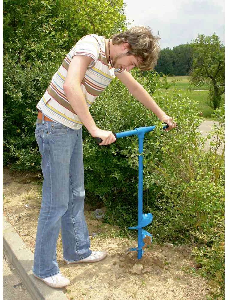 Eine Person bohrt mit einem blauen Handerdbohrer ein Loch im Boden neben Büschen.