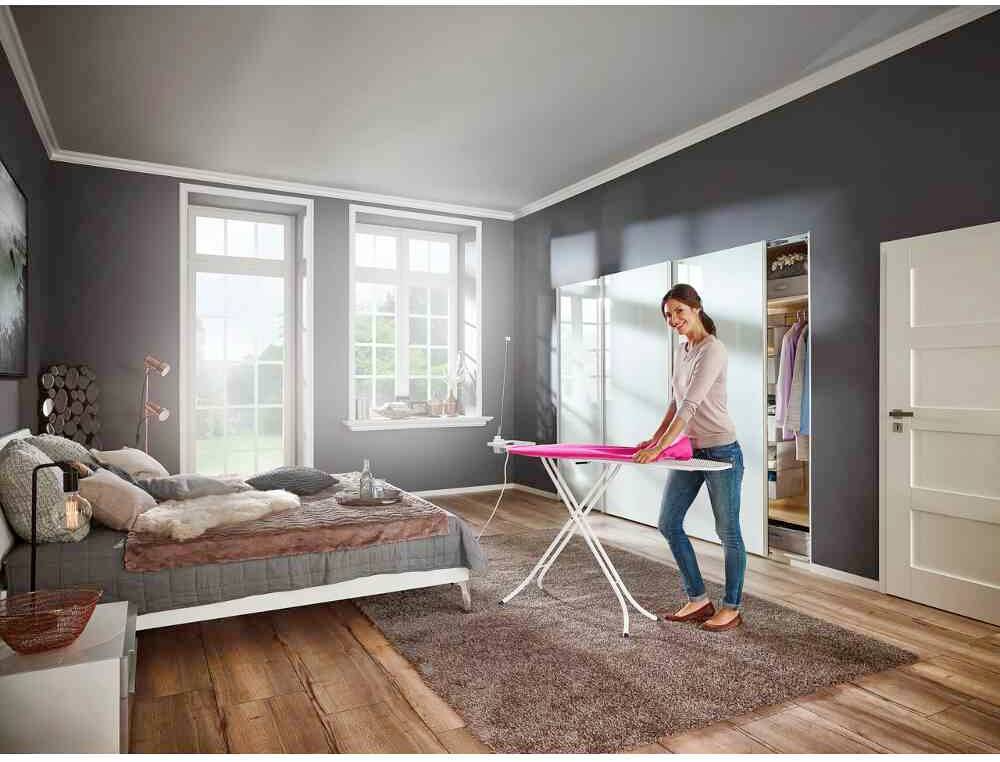 Eine Frau bügelt Kleidung in einem Schlafzimmer mit einem Bett und großen Fenstern. Die Atmosphäre wirkt hell und gemütlich.