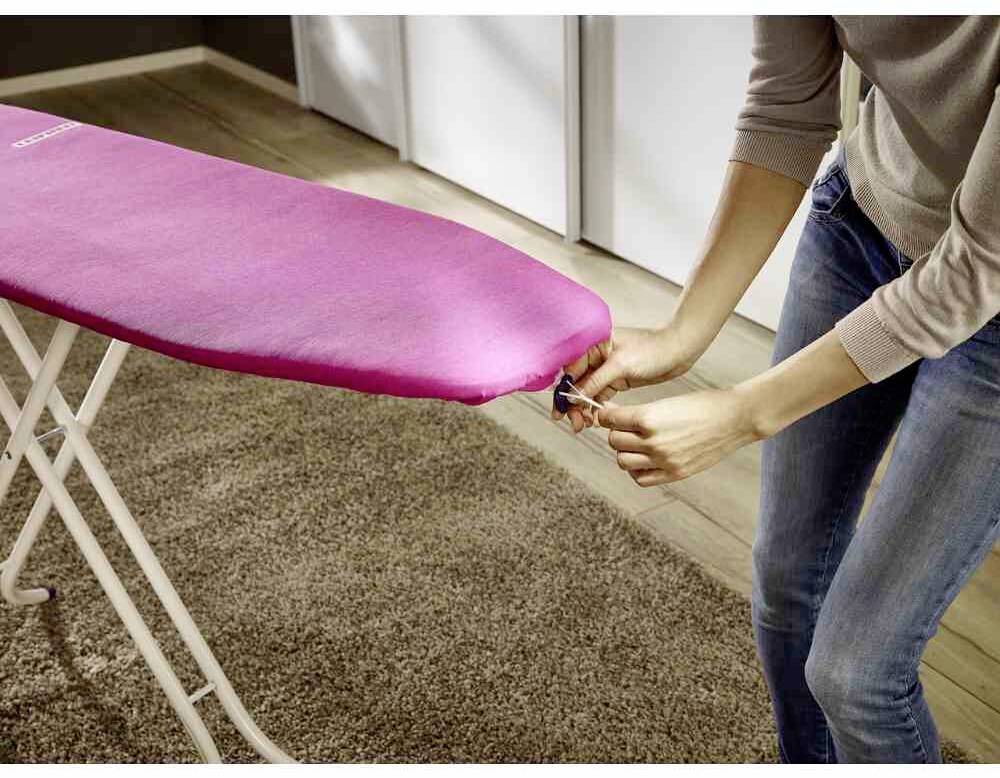 Person zieht den Bezug auf einem Bügelbrett in einem Innenraum fest. Das Bügelbrett hat eine pinke Abdeckung und steht auf einem Teppich.