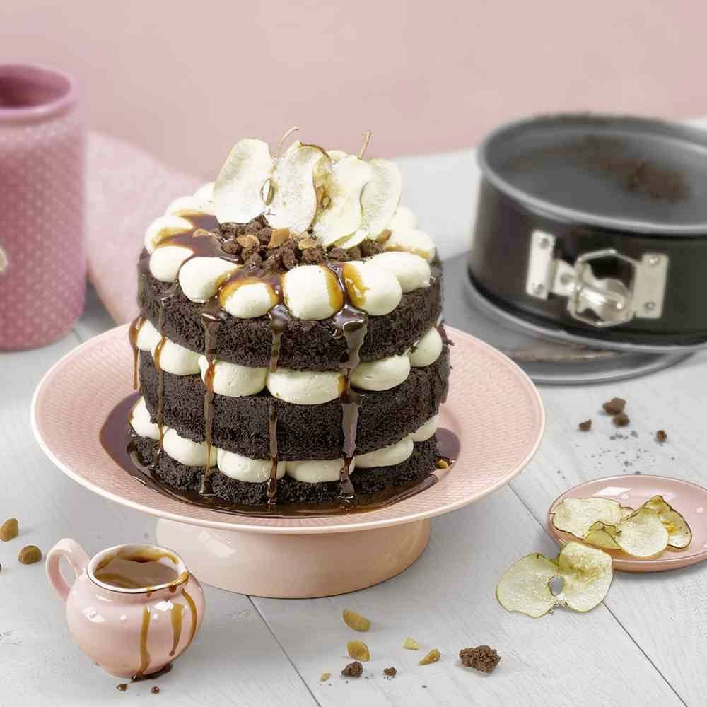 Schokoladenschichtkuchen mit Sahne und Karamellsauce, dekoriert mit Apfelscheiben auf rosa Teller. Hintergrund enthält Backzubehör.