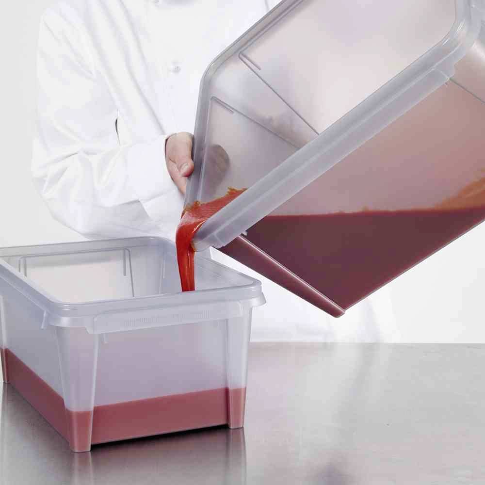 Ein Koch in weißem Kittel gießt eine rote Flüssigkeit von einem durchsichtigen Behälter in einen anderen auf einer Metalloberfläche.