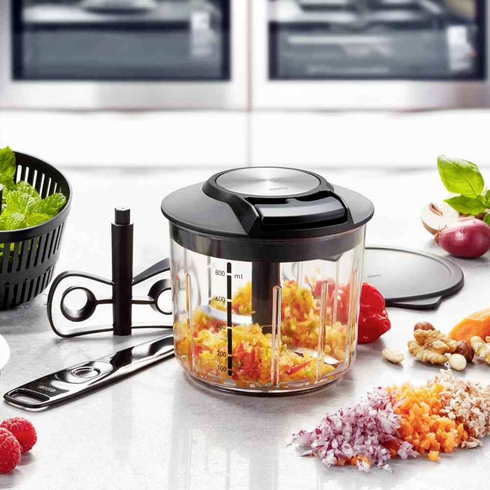 Küchenmaschine auf Tisch, gefüllt mit zerkleinertem Gemüse. Im Hintergrund liegen Zutaten wie Kräuter, Zwiebeln und Nüsse.