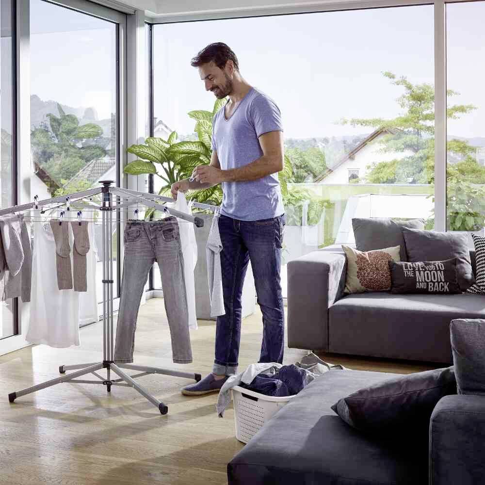 Ein Mann hängt Kleidung im Wohnzimmer auf einem Wäscheständer zum Trocknen auf. Im Hintergrund Fenster mit Blick ins Freie.