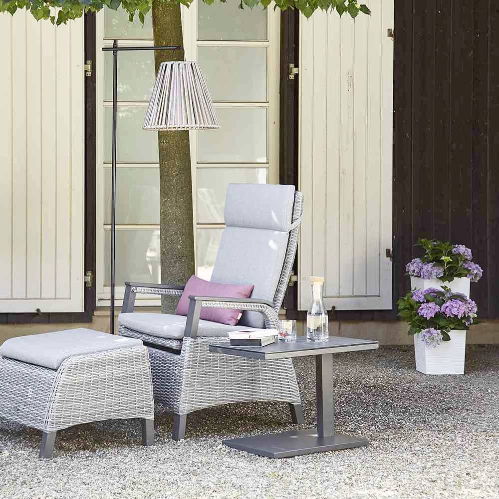Ein grauer Rattan-Loungesessel mit Hocker unter einem Baum. Daneben ein kleiner Tisch mit Karaffe und Blume. Im Hintergrund helle Türen.
