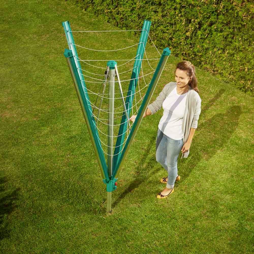 Eine Frau steht lächelnd neben einem grünen Wäscheschirm in einem Garten. Der Schirm hat ausgebreitete Leinen zum Trocknen von Kleidung.