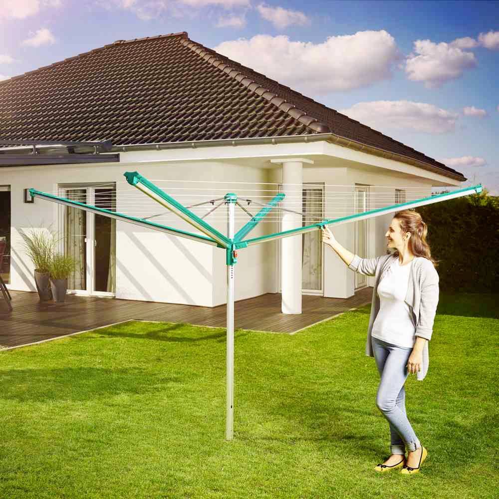 Eine Frau steht neben einer modernen Wäschespinne im Garten vor einem Einfamilienhaus. Der Himmel ist leicht bewölkt.