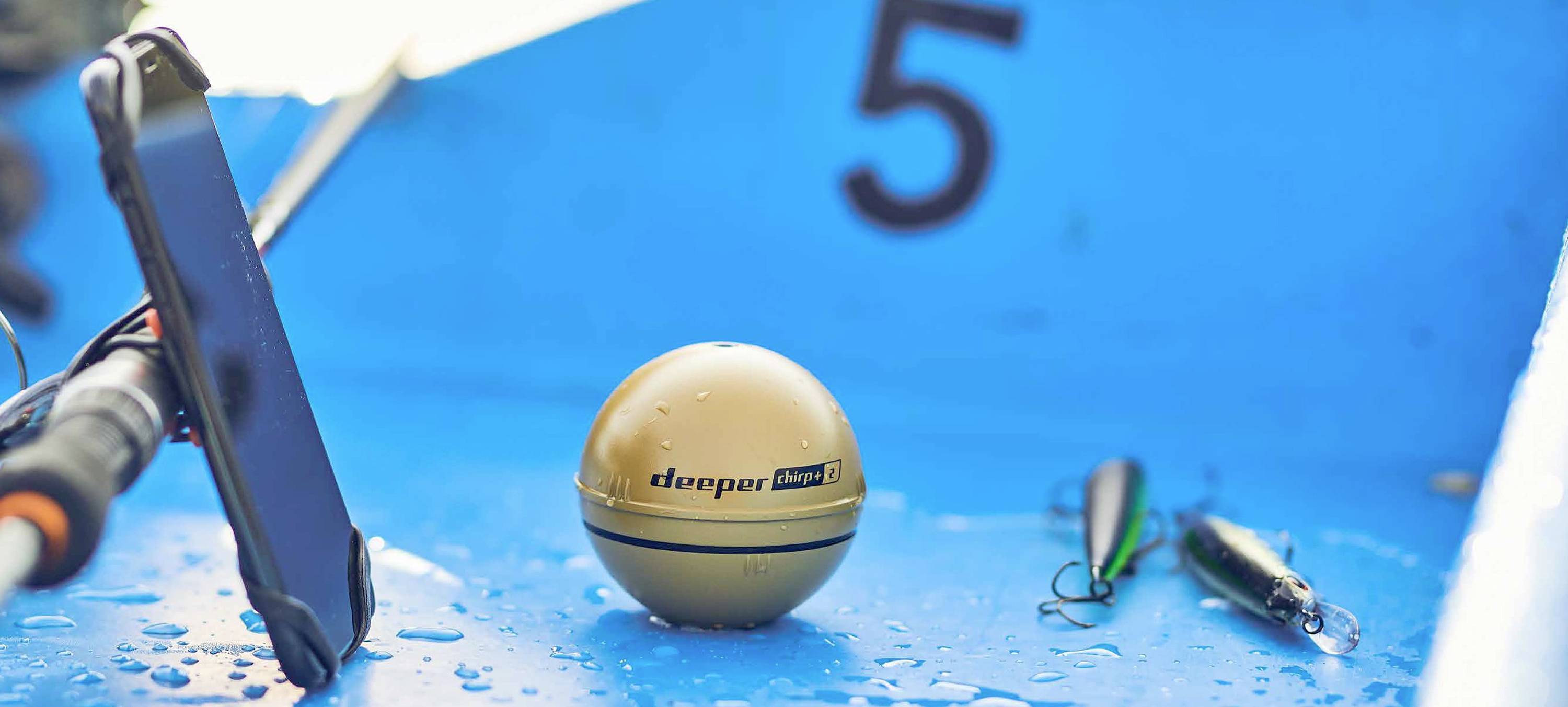 Ein goldener Sonarball mit der Aufschrift 'deeper' liegt auf einem blauen Boot neben einem Smartphone und einem Angelköder.