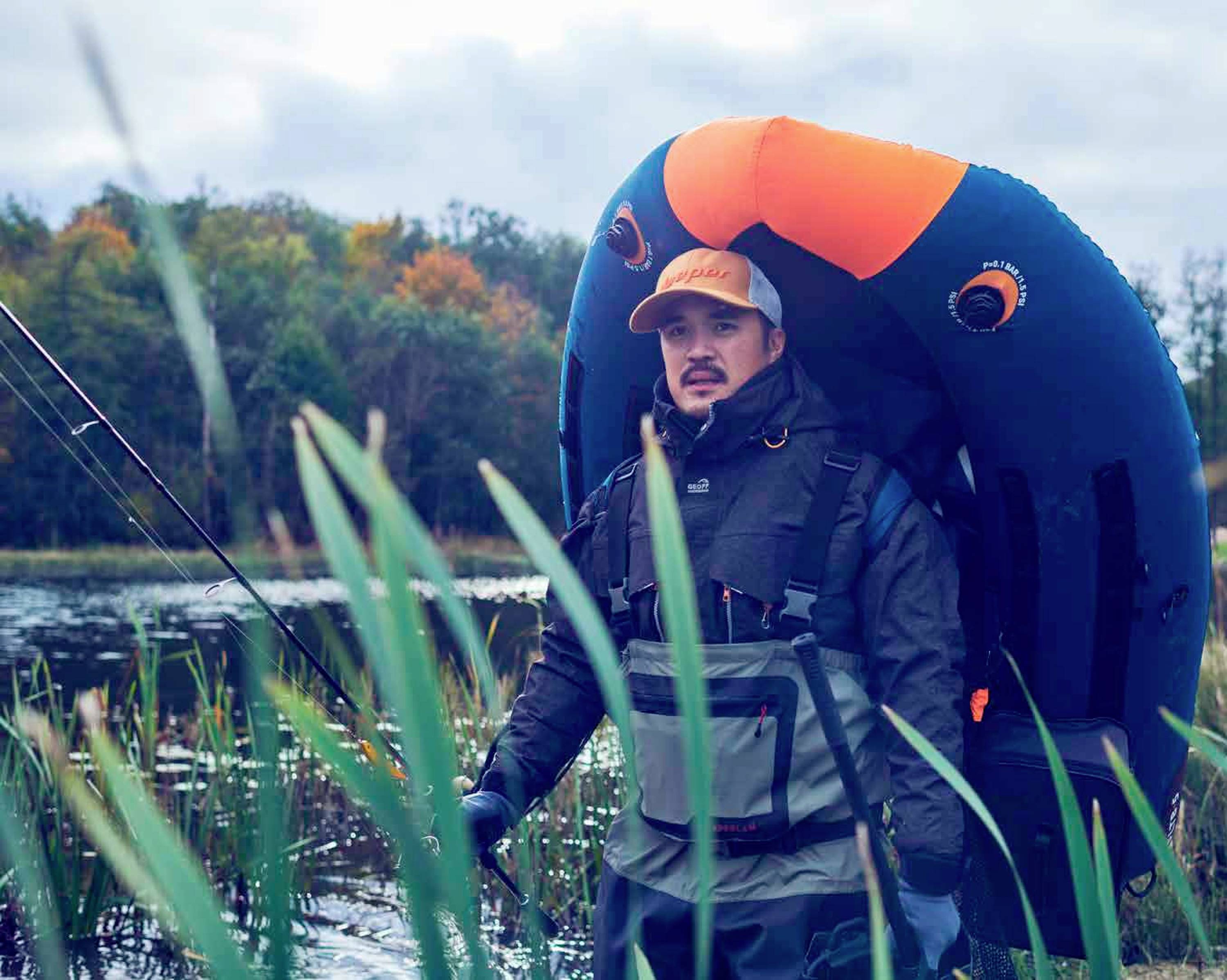 Mann in Anglerbekleidung steht mit einer aufblasbaren Schwimmhilfe auf dem Rücken in einem See mit Schilf und Bäumen im Hintergrund.