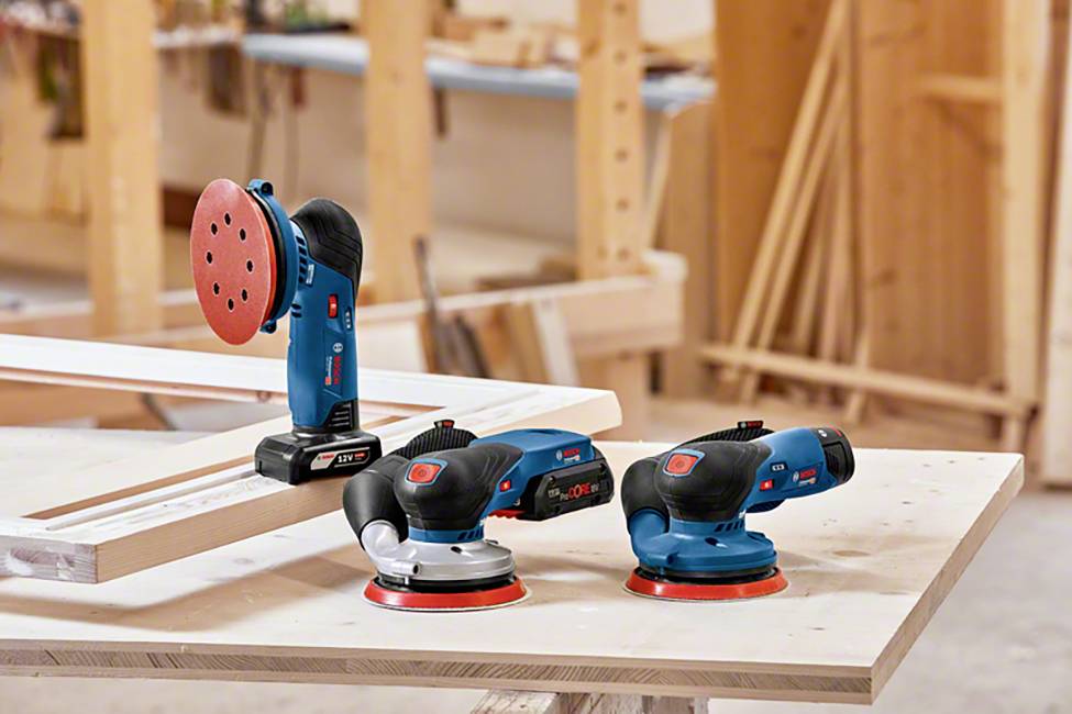 Drei Schleifmaschinen auf einem Holztisch in einer Werkstatt. Die Werkzeuge haben rote und blaue Elemente. Hintergrund zeigt Holzregale.