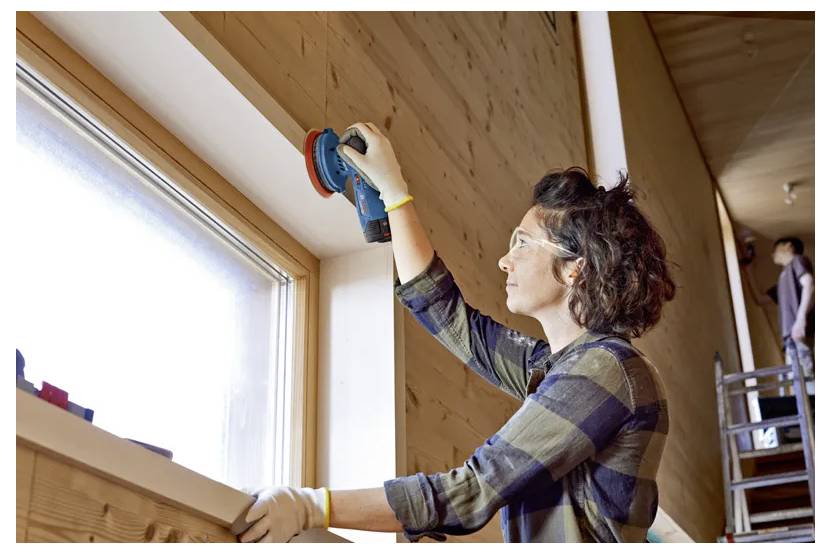 Eine Person, die einen Bandschleifer an einem Holzfensterrahmen in einem gut beleuchteten Raum verwendet. Sie trägt Schutzbrille und Handschuhe.
