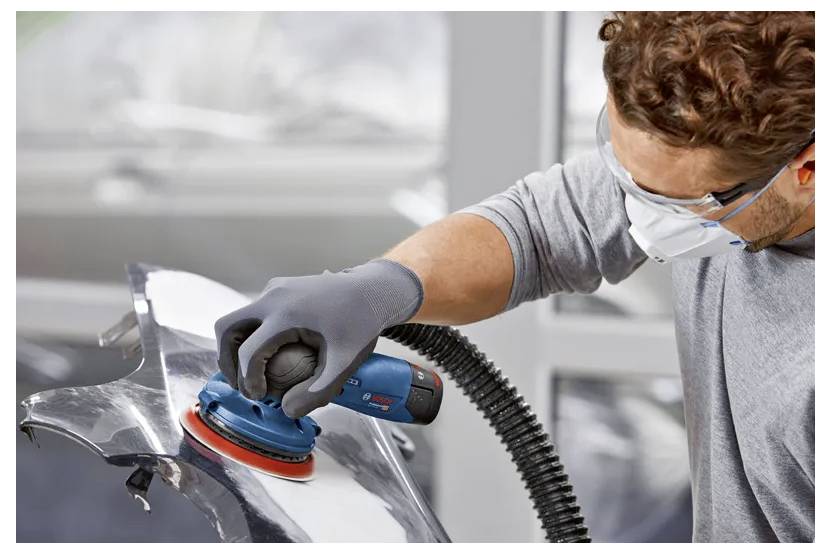 Eine Person mit Schutzbrille und Maske benutzt einen Bandschleifer auf einer Metalloberfläche und sorgt für eine glatte Oberfläche in einer Werkstattumgebung.