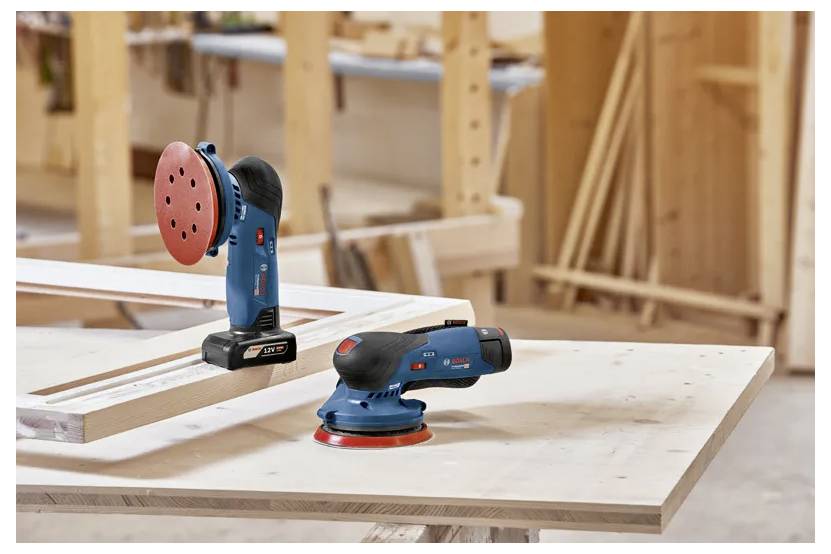 Zwei kabellose Schleifmaschinen auf einer Werkbank in einer Holzwerkstatt, mit Holzregalen und Werkzeugen im Hintergrund.