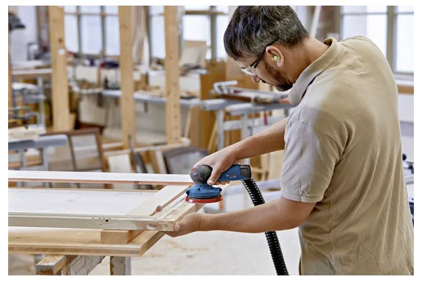Ein Mann mit Gehörschutz benutzt einen Exzenterschleifer an einem Holzrahmen in einer Werkstatt, die mit Holzbearbeitungswerkzeugen und -materialien gefüllt ist.
