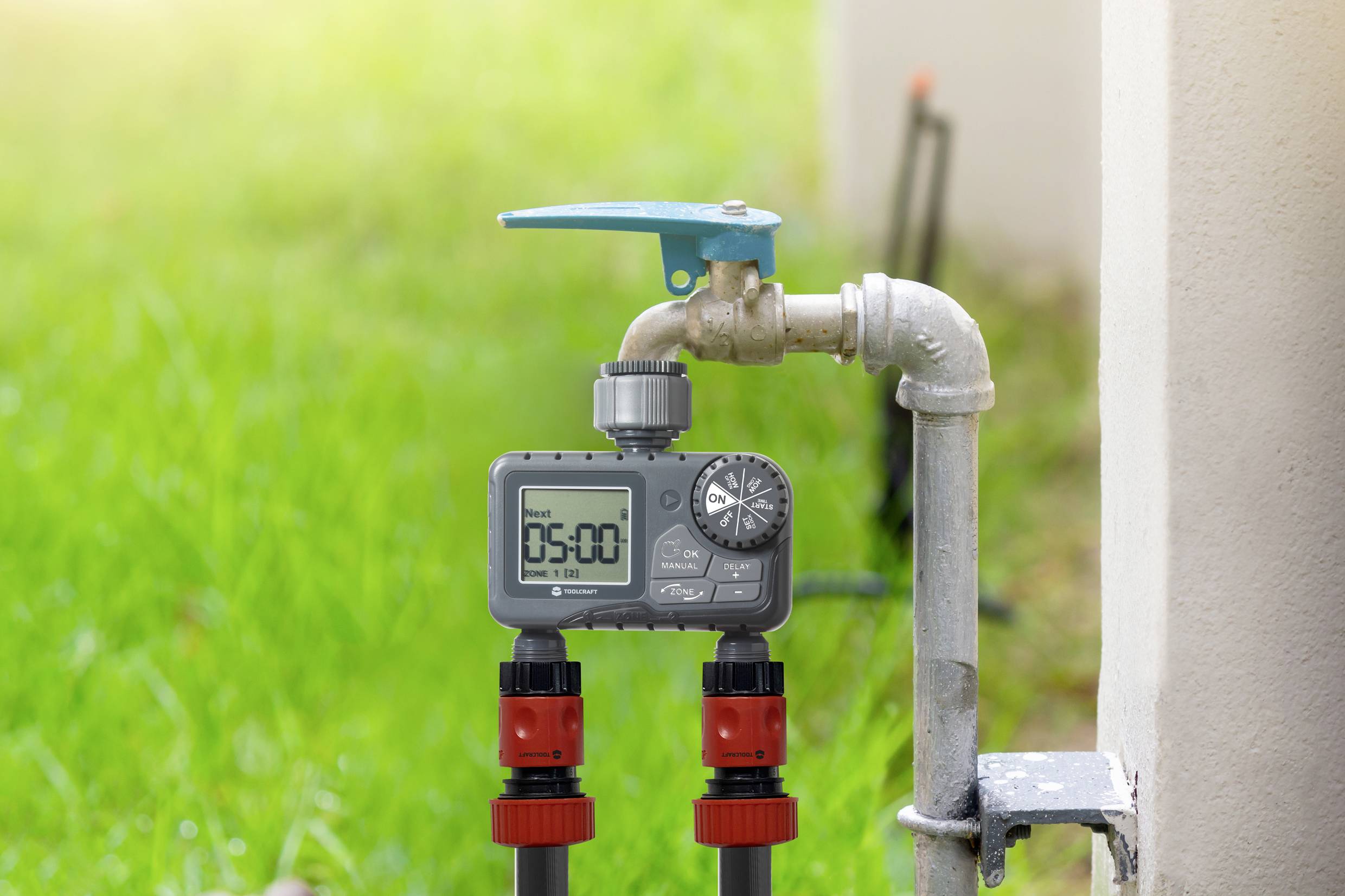 Ein digitaler Bewässerungstimer ist an einem Wasserhahn im Freien befestigt. Der Timer zeigt '05:00' an. Im Hintergrund ist Gras zu sehen.