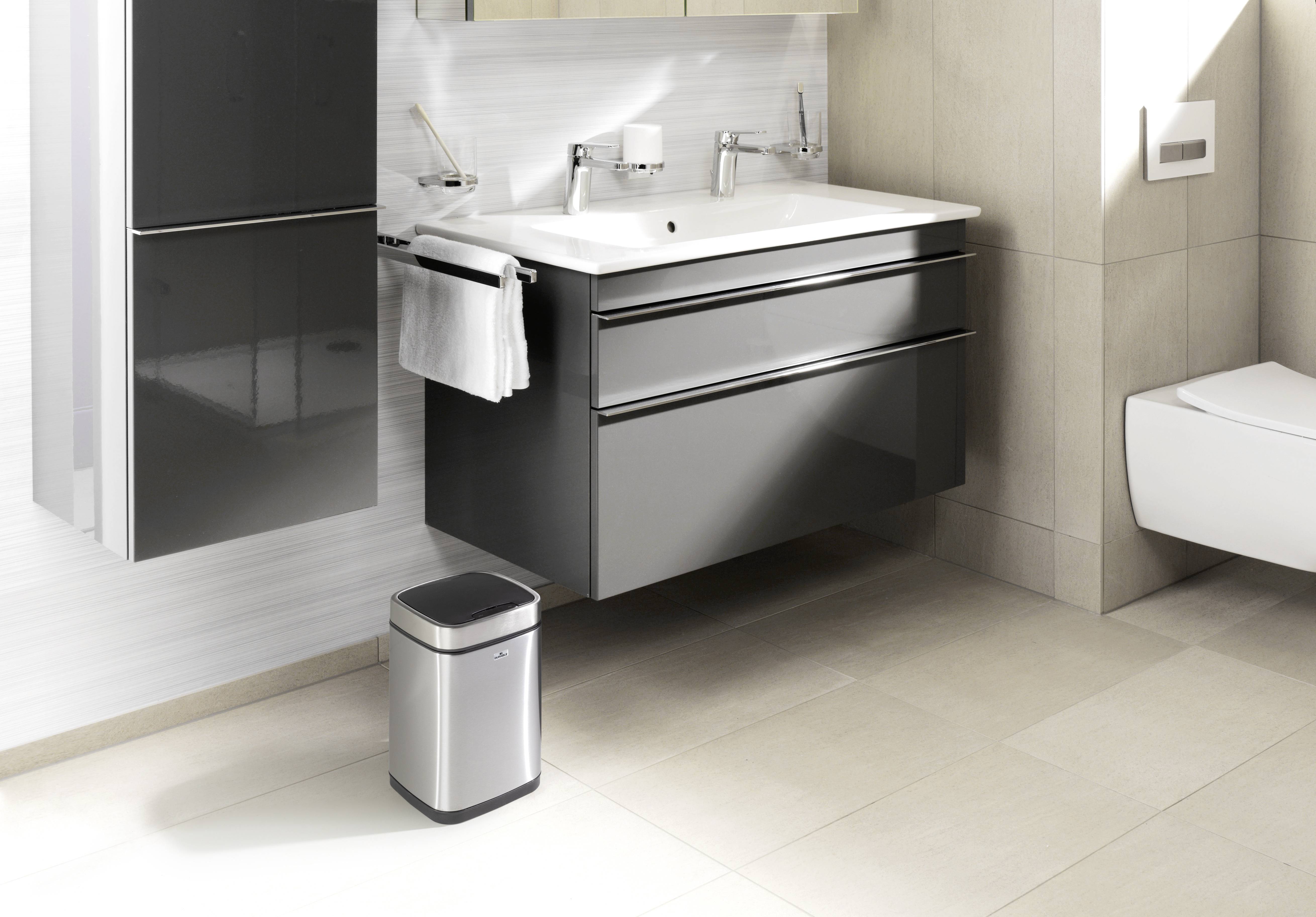 Modernes Badezimmer mit zwei Waschbecken, schwarzem Unterschrank, Handtuchhalter und kleinem Mülleimer auf beigem Fliesenboden.