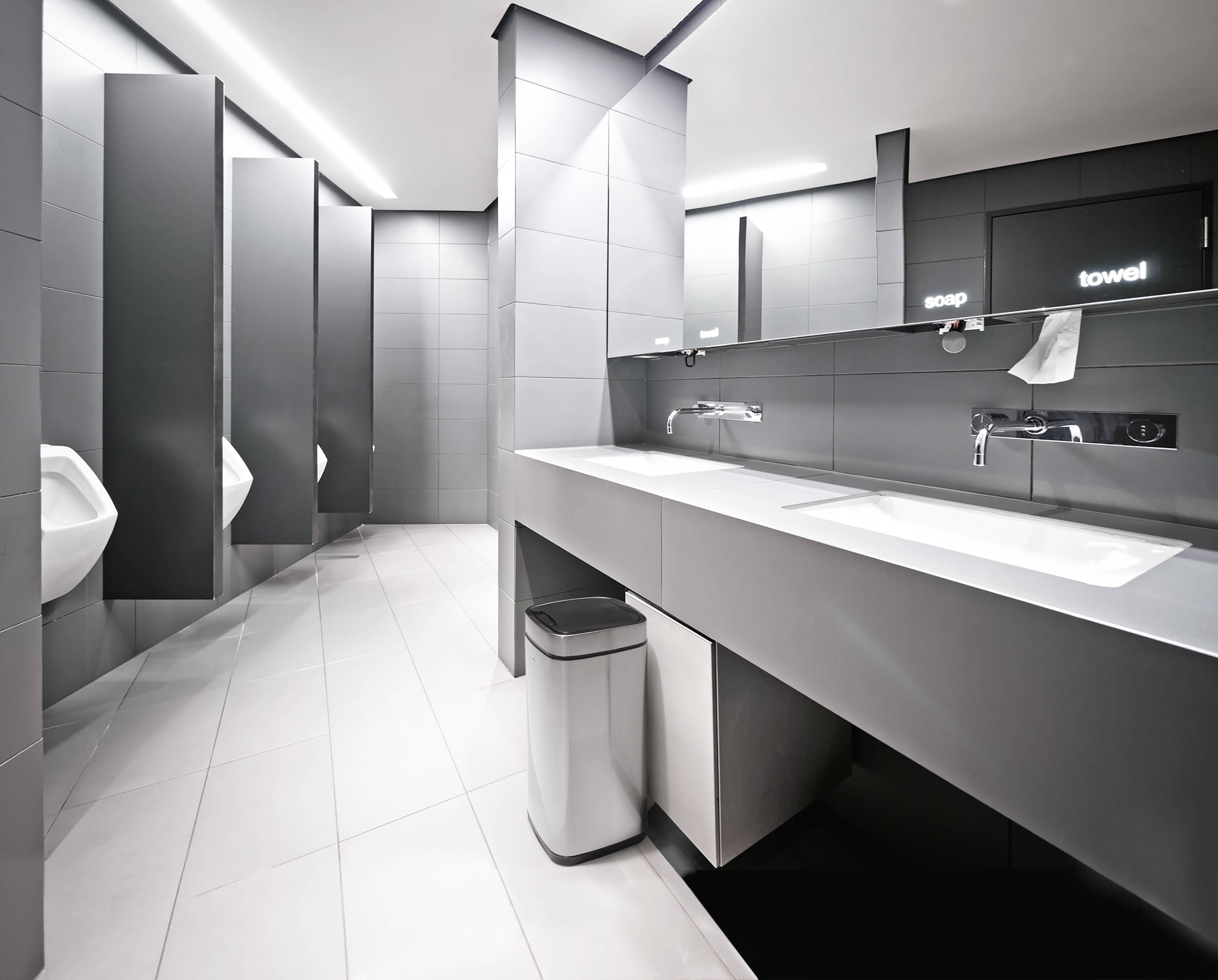 Öffentliche Männertoilette mit moderner Ausstattung, einschließlich Waschbecken, Spiegel, Urinale und Mülleimer in einem sauberen, grauen Design.