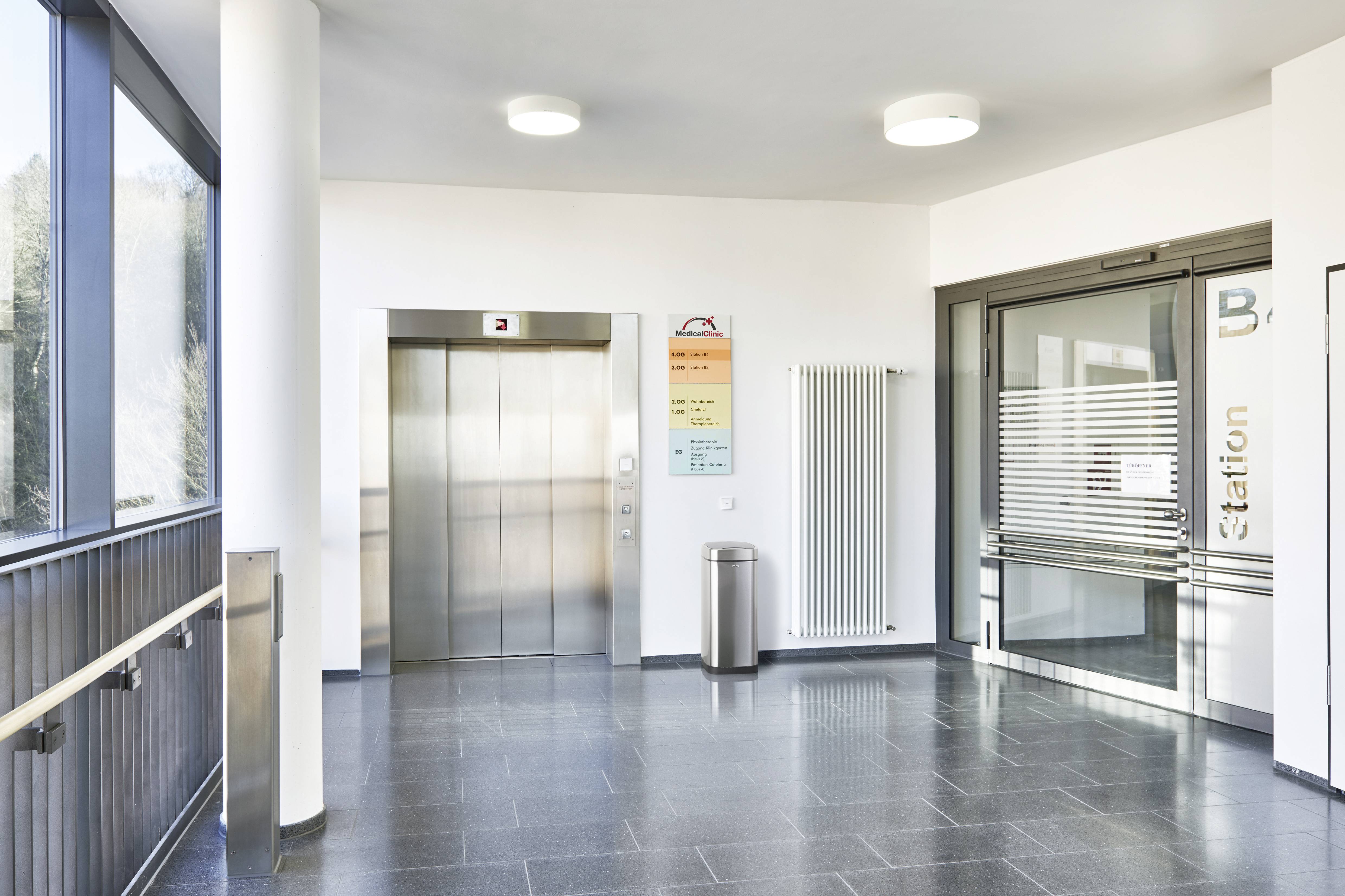 Ein heller Krankenhausflur mit einem Fahrstuhl auf der linken Seite und einer Glastür zur Station B rechts. Ein Abfalleimer steht neben dem Fahrstuhl.